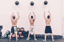 Vista posteriore di tre cross trainer maschi che lanciano palle da ginnastica in palestra — Foto stock
