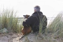 Задній вид людини і ПЕТ собака, сидячи на піщані дюни, Костянтин Bay, Корнуолл, Великобританія — стокове фото