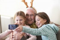 Sisters taking smartphone selfie with grandfather in reindeer antlers — Stock Photo