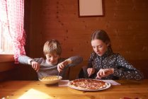 Девочка-подросток и брат едят макароны и пиццу за столом в шале — стоковое фото
