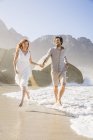 Полный вид спереди пары, бегущей по пляжу, держась за руки — стоковое фото