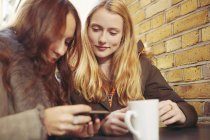 Zwei Freundinnen, die draußen sitzen, Kaffee trinken und aufs Smartphone schauen — Stockfoto