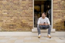 Hombre joven con teléfono inteligente sentado en la puerta, Kings Road, Londres, Reino Unido - foto de stock