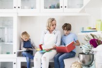Femme mûre assise sur le comptoir de cuisine lisant des livres d'histoires avec fils et fille — Photo de stock