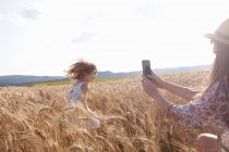 Madre fotografiando chica corriendo a través del campo de trigo - foto de stock