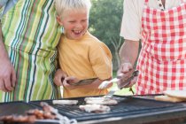 Мальчик готовит шашлыки и бургеры на барбекю с отцом и дедушкой — стоковое фото