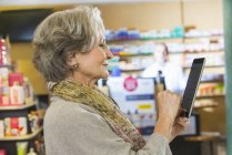 Старшая женщина, использующая цифровой планшет для проверки лекарств онлайн в аптеке — стоковое фото