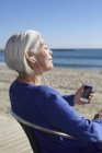 Reife Frau hört Musik mit Ohrhörern am Strand — Stockfoto
