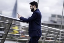 Homme d'affaires debout sur la passerelle lisant le texte d'un smartphone, Londres, Royaume-Uni — Photo de stock