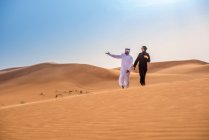 Couple wearing traditional middle eastern clothes pointing from desert dune, Dubai, United Arab Emirates — Stock Photo