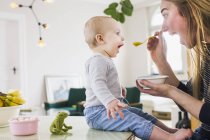 Bebê menina mimetizando mãe enquanto come na mesa da cozinha — Fotografia de Stock