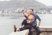 Casal jovem sentado na parede do porto tomando selfie smartphone, Lago de Como, Itália — Fotografia de Stock