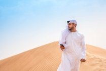 Homme du Moyen-Orient portant des vêtements traditionnels à l'aide d'un smartphone sur une dune du désert, Dubaï, Émirats arabes unis — Photo de stock