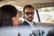 Joven hombre de negocios y mujer hablando en el asiento trasero del coche, Dubai, Emiratos Árabes Unidos - foto de stock