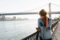 Mujer joven usando teléfono celular, Manhattan Bridge, Brooklyn, EE.UU. - foto de stock