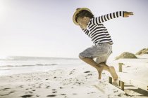 Jeune garçon sautant sur la plage, portant un chapeau de paille — Photo de stock