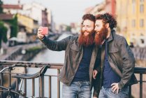 Молодые хипстеры-близнецы с рыжими волосами и бородами делают селфи на берегу канала — стоковое фото