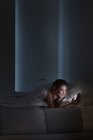 Mujer joven acostada en la cama leyendo textos de teléfonos inteligentes por la noche - foto de stock