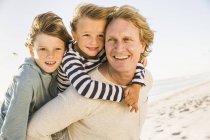 Сини на пляжі з обіймами навколо батька, дивлячись в очі посміхаючись — стокове фото