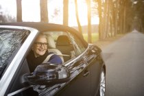 Жінка в машині, що їде на дереві викладена дорога дивиться з вікна посміхаючись — стокове фото