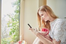 Jovem mulher lendo texto do smartphone na cozinha — Fotografia de Stock