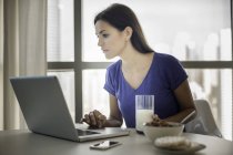 Jovem mulher usando laptop enquanto toma café da manhã — Fotografia de Stock