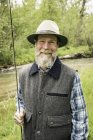 Бородатий чоловік тримає вудку на річці, дивлячись на камеру посміхаючись — стокове фото
