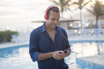 Взрослый мужчина, выбирающий музыку со смартфона у бассейна отеля, Рио-де-Жанейро, Бразилия — стоковое фото