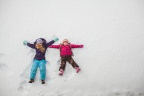 Две сестры играют, делая снежных ангелов в снегу — стоковое фото