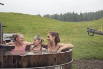 Tres amigas riendo mientras se relajan en la bañera de hidromasaje rural, Sattelbergalm, Tirol, Austria - foto de stock