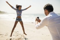 Homme mûr photographiant sa petite amie sautant, plage d'Arpoador, Rio De Janeiro, Brésil — Photo de stock