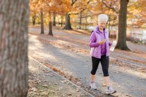 Senior woman walking outdoors, using smartphone, wearing earphones and holding water bottle — Stock Photo