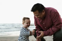 Отец на пляже с маленьким мальчиком, смотрящим на камень — стоковое фото