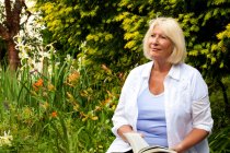 Senior mulher sentada no jardim segurando livro — Fotografia de Stock