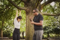 Entrenador personal instruyendo a la mujer en pull-ups utilizando la rama del árbol del parque - foto de stock