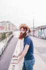 Young male hipster with red hair and beard listening to headphones on city bridge — Stock Photo