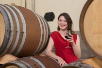 Portrait of young woman in wine cellar, leaning against wine barrel, holding glass of wine — Stock Photo