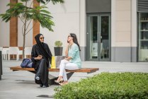 Joven mujer de mediana edad vestida con ropa tradicional sentada en el banquillo con una amiga, Dubai, Emiratos Árabes Unidos. - foto de stock