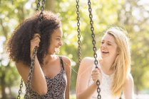 Dos mujeres jóvenes sentadas en el parque columpio riendo - foto de stock
