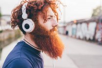 Young male hipster with red hair and beard listening to headphones in city — Stock Photo