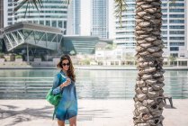 Turista femenina paseando en textos de lectura de teléfonos inteligentes frente al mar, Dubai, Emiratos Árabes Unidos - foto de stock