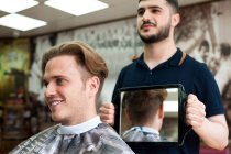 Peluquero usando espejo para mostrar el corte de pelo sonriente del cliente - foto de stock