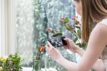Над видом на плече жінки, використовуючи смартфон, щоб сфотографувати квітку у вазі — стокове фото