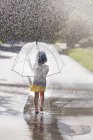 Vista trasera de chica descalza llevando paraguas caminando por el charco de la calle - foto de stock