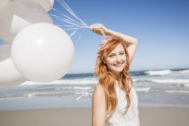 Rothaarige Frau am Strand hält Luftballons in die Kamera und lächelt — Stockfoto