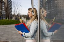 Красива блондинка студентка, що стоїть у скляному вікні, тримає мобільний телефон і папки, носить навушники — стокове фото