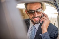 Jeune homme d'affaires portant des lunettes de soleil parlant sur un smartphone dans un siège arrière de voiture, Dubaï, Émirats arabes unis — Photo de stock