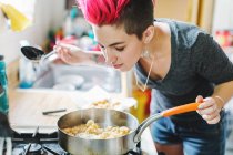 Молодая женщина с розовыми волосами нюхает жареную пищу на кухонной плите — стоковое фото
