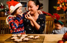 Madre y dos hijos en la mesa comiendo galletas de Navidad caseras - foto de stock