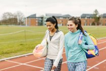 Dos mujeres jóvenes caminando en pista de atletismo, llevando bolsas deportivas - foto de stock